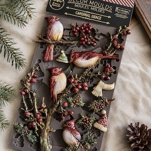 May include: A gray silicone decor mould titled Cardinal Grace featuring cavities for four cardinal birds perched on branches with berries, pinecones, a scarf, and a winter hat. The packaging header includes the text Decor Moulds and re-design.