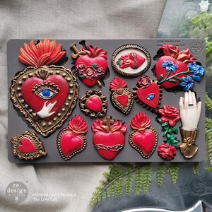 May include: A gray silicone mold filled with various red heart-shaped designs, including flaming hearts, hearts with crosses, and hearts with floral accents. Some hearts have gold detailing and a hand with a ring. The text "re design with prima" is visible.