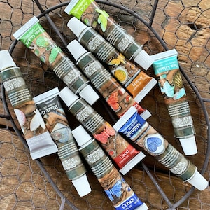 May include: An assortment of Art Alchemy Metallique Wax tubes in various colors, including Moss Green, Black Gold, and Cobalt. Each tube features decorative artwork and color names. The tubes are arranged on a wire mesh surface.