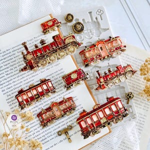 May include: A collection of red and gold metal train and trolley charms arranged on a white silicone mold. The set includes various locomotive and carriage designs with intricate wheel details, alongside small metal key-shaped embellishments.