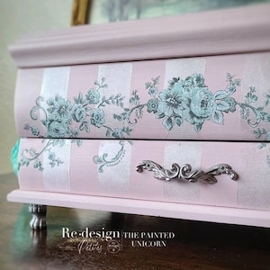 May include: A pink jewelry box with silver stripes and floral accents. The box features a decorative silver handle and rests on small, ornate feet. The text "Re-design Petites" and "The Painted Unicorn" is visible.