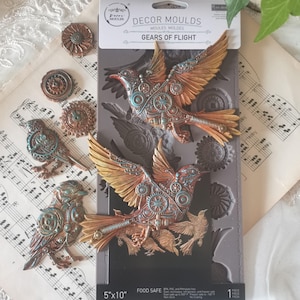 May include: A silicone mold for making decorative bird-shaped cake toppers. The mold features two birds with intricate steampunk designs, including gears and cogs. The mold is 5 inches by 10 inches and is food safe.