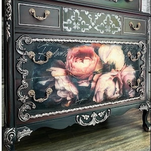 May include: A dark-coloured chest of drawers with elaborate silver and gold detailing. The drawers display a floral design with pink and white roses. The top drawers have a patterned design and gold handles. The word "Kindness" is written on the floral drawer.