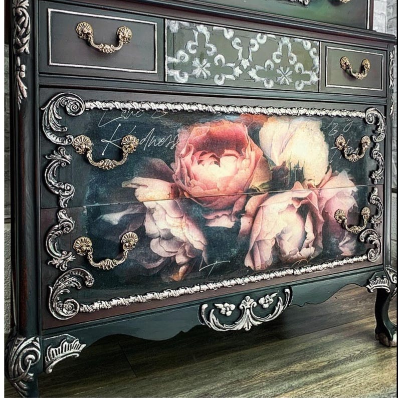May include: A dark-coloured chest of drawers with elaborate silver and gold detailing. The drawers display a floral design with pink and white roses. The top drawers have a patterned design and gold handles. The word "Kindness" is written on the floral drawer.