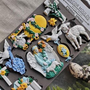 May include: A gray silicone mold labeled ETERNAL SPRING, ITEM NO 681715, featuring cavities for casting shapes including a lamb, an angel with a lamb, doves, a yellow egg with flowers, a cross with lilies, and various floral arrangements.