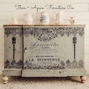 Op de afbeelding: Een lichtgrijs houten dressoir met zwarte kantachtige patronen en Franse typografie, waaronder "Sp&eacute;cialit&eacute;s du menu" en "LA BIENVENUE". De kast heeft een blad van natuurlijk hout, een kleine witte knop en vijf witte keramische kannen.