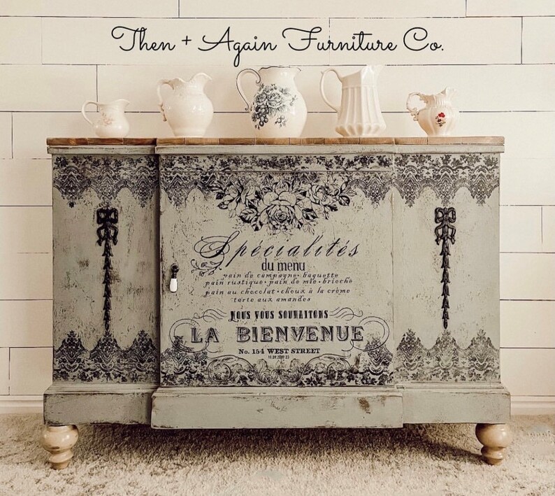 Op de afbeelding: Een lichtgrijs houten dressoir met zwarte kantachtige patronen en Franse typografie, waaronder "Sp&eacute;cialit&eacute;s du menu" en "LA BIENVENUE". De kast heeft een blad van natuurlijk hout, een kleine witte knop en vijf witte keramische kannen.