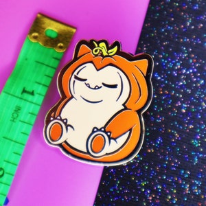 Op de afbeelding: Een gouden emaille pin met een cartoonfiguur dat een oranje pompoenkostuum draagt. Het personage heeft gesloten ogen en een glimlach. De pin is op een roze en zwarte glitterachtergrond.