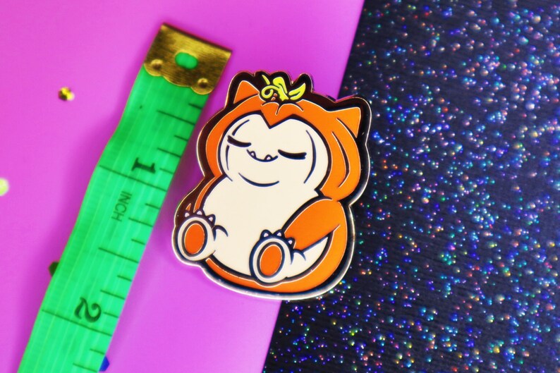 Op de afbeelding: Een gouden emaille pin met een cartoonfiguur dat een oranje pompoenkostuum draagt. Het personage heeft gesloten ogen en een glimlach. De pin is op een roze en zwarte glitterachtergrond.
