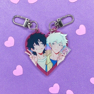May include: A heart-shaped keychain with two anime characters on it. The characters are a young man with black hair and a young man with white hair. The keychain is pink and has a silver chain.
