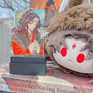 May include: A colorful enamel pin featuring a character holding a flower, set against a blue sky and sun. The pin is on a black stand, with a plush toy in the background. The pin has red, brown, and white accents.