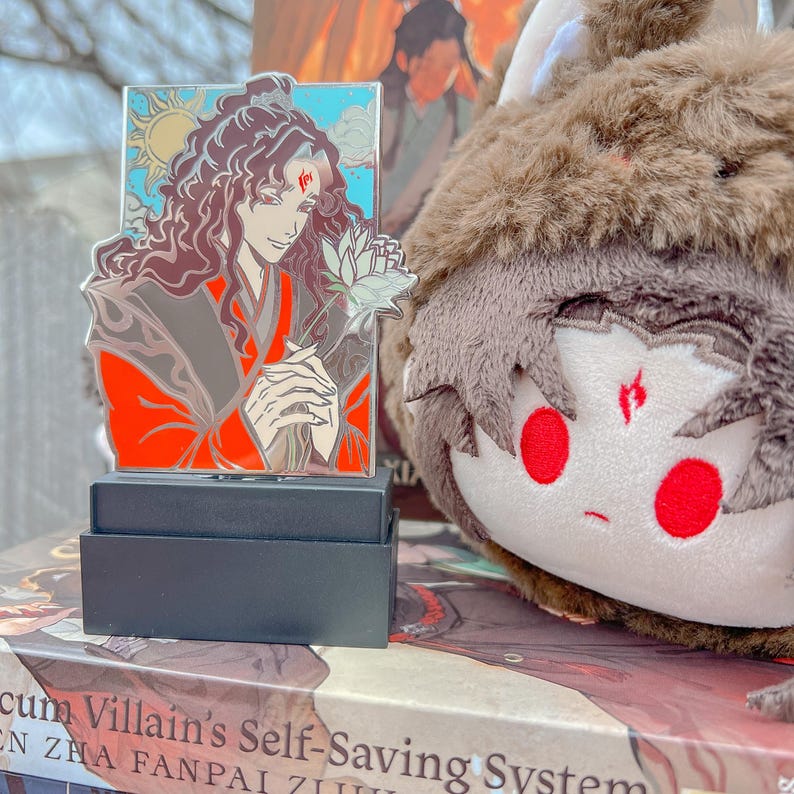 May include: A colorful enamel pin featuring a character holding a flower, set against a blue sky and sun. The pin is on a black stand, with a plush toy in the background. The pin has red, brown, and white accents.