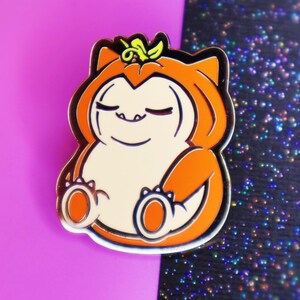 Op de afbeelding: Een gouden emaille pin met een cartoonfiguur dat een pompoenkostuum draagt. Het figuur heeft een rond, oranje lichaam met een wit gezicht en gesloten ogen. Het figuur draagt een bruine hoed met een groene steel erop.