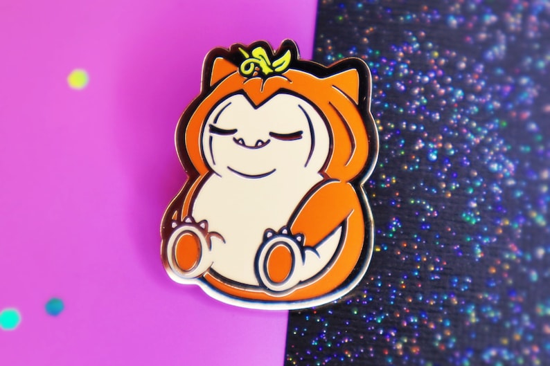 Op de afbeelding: Een gouden emaille pin met een cartoonfiguur dat een pompoenkostuum draagt. Het figuur heeft een rond, oranje lichaam met een wit gezicht en gesloten ogen. Het figuur draagt een bruine hoed met een groene steel erop.