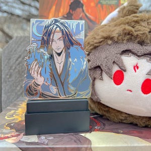 May include: A detailed enamel pin featuring a character with long dark hair and blue clothing, set against a backdrop of a crescent moon and icicles. The pin is displayed on a black box, with a plush toy in the background.