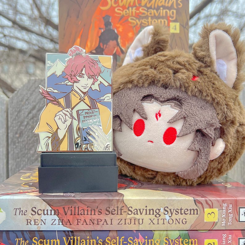 May include: A colorful enamel pin featuring a character with pink hair, holding a quill and a book with the text "PROUD IMMORTAL DEMON WAY." A plush toy with a brown furry hat and red eyes sits next to the pin. Books with the title "The Scum Villain's Self-Saving System" are in the background.