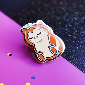 Op de afbeelding: Een gouden emaille pin met een cartoonfiguur met oranje vacht, gesloten ogen en een kroon. Het figuur is opgerold in een bal, met zijn poten zichtbaar.