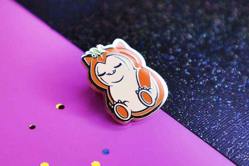 Op de afbeelding: Een gouden emaille pin met een cartoonfiguur met oranje vacht, gesloten ogen en een kroon. Het figuur is opgerold in een bal, met zijn poten zichtbaar.