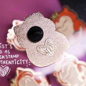 Op de afbeelding: De achterkant van een gouden emaille pin met een cartoonfiguur. De pin heeft een zwarte rubberen achterkant en een hartvormig logo met de tekst "Downright Sketchy".