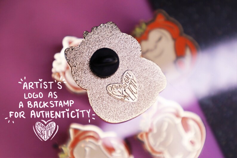 Op de afbeelding: De achterkant van een gouden emaille pin met een cartoonfiguur. De pin heeft een zwarte rubberen achterkant en een hartvormig logo met de tekst "Downright Sketchy".