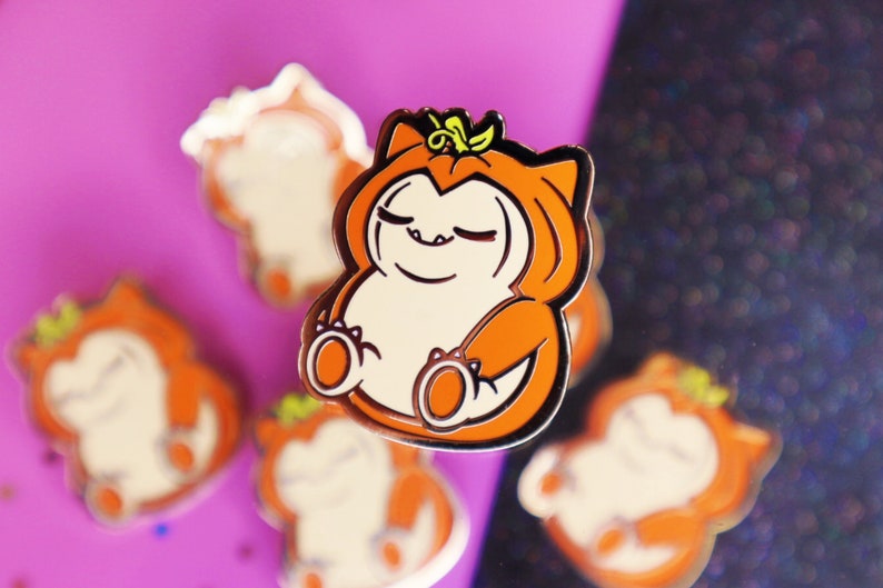 Op de afbeelding: Een set van vijf oranje en witte emaille pinnen met een cartoonfiguur dat verkleed is als een pompoen. Het personage heeft een glimlach met gesloten ogen en draagt een pompoenkostuum met een groene steel erop.