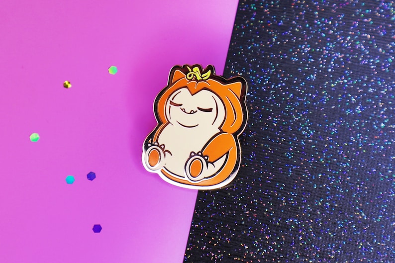 Op de afbeelding: Een gouden emaille pin met een cartoonfiguur dat een oranje en bruin pompoenkostuum draagt. Het personage heeft zijn ogen dicht en een ontspannen uitdrukking.