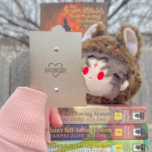 May include: A silver-colored pin with the text "IVYINSIDE" and a heart design. A plush toy with a brown bear hat and red eyes is visible. Books with the title "The Scum Villain's Self-Saving System" are stacked in the background.