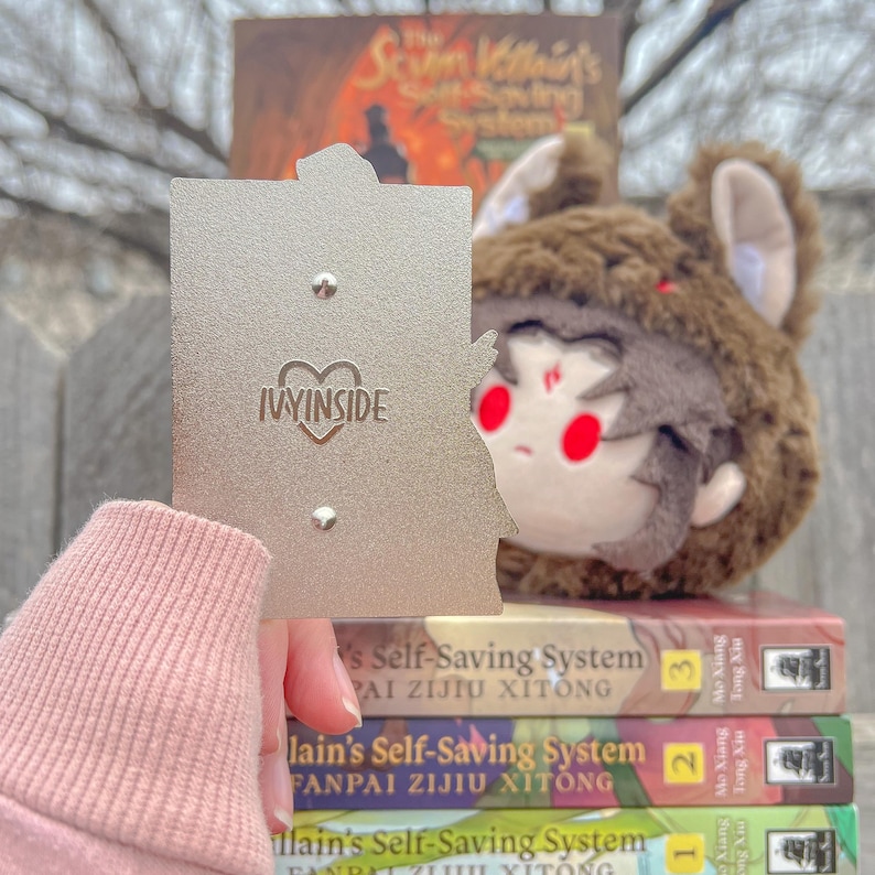 May include: A silver-colored pin with the text "IVYINSIDE" and a heart design. A plush toy with a brown bear hat and red eyes is visible. Books with the title "The Scum Villain's Self-Saving System" are stacked in the background.