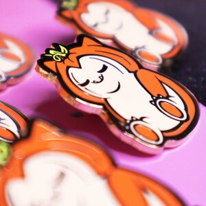 Op de afbeelding: Close-up van een set oranje en witte emaillepins met een cartoonkat die een kroon draagt. De pins hebben een gouden afwerking en zijn gerangschikt op een roze oppervlak.