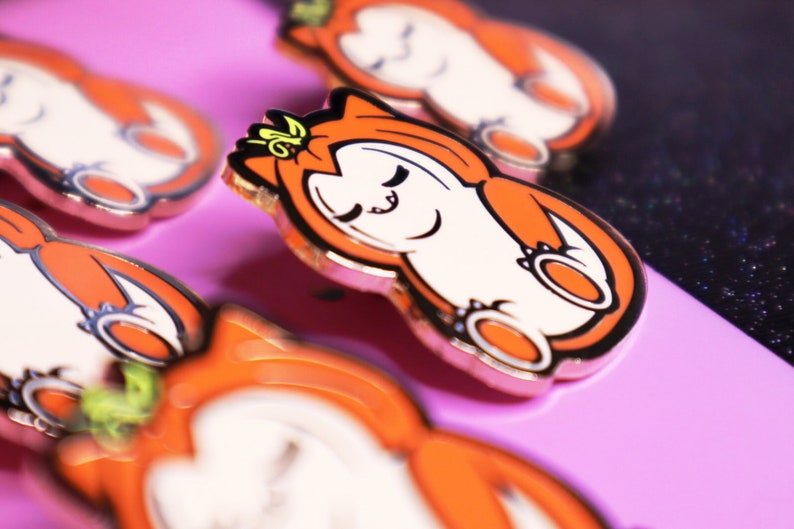 Op de afbeelding: Close-up van een set oranje en witte emaillepins met een cartoonkat die een kroon draagt. De pins hebben een gouden afwerking en zijn gerangschikt op een roze oppervlak.
