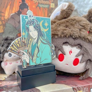 May include: A teal and gold enamel pin featuring a character with long black hair, holding a fan. The pin is set against a blue background with a crescent moon and stars. It is displayed on a black stand.