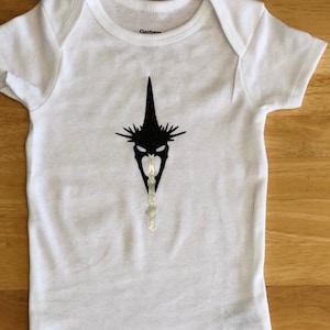 May include: White baby onesie with a black and white graphic of a character from the Lord of the Rings. The graphic is of the Nazgûl, a ringwraith, with a pointed hat and a menacing face.