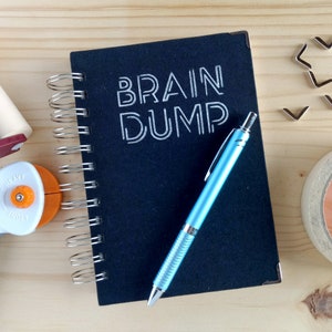 Może przedstawiać: Czarny notes z spiralą, na okładce napis "BRAIN DUMP" w kolorze białym. Na notesie leży niebieski długopis.