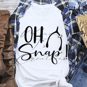 May include: White t-shirt with the text "Oh Snap!" in black cursive lettering and a horseshoe graphic. The shirt is laid out on a wooden surface with a pair of brown boots, a blue plaid shirt, and a straw cowboy hat.