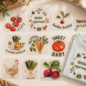 May include: A collection of iron-on transfers featuring watercolor illustrations of vegetables, a chicken, and text including "little sprout," "hello baby," "locally grown," "freshly picked," "garden baby," "sweet baby," and "homegrown with love."
