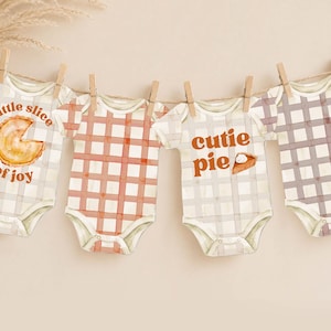 Puede incluir: Cuatro bodies de bebé colgados de una cuerda con pinzas de madera. Dos tienen un patrón de cuadros en naranja y gris, mientras que los otros muestran gráficos de tartas con el texto "little slice of joy" y "cutie pie" en letras naranjas.