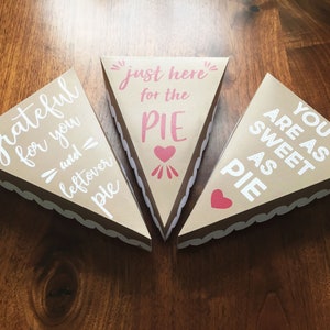 Op de afbeelding: Drie bruine papieren taartdozen met witte gegolfde randen. De dozen zijn versierd met witte tekst en roze harten. De tekst op de dozen luidt "Grateful for you and leftover pie", "Just here for the pie" en "You are as sweet as pie".