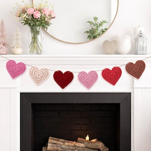 May include: A decorative garland featuring seven crocheted hearts in various shades of pink, red, and brown, each with a white border, strung on a brown cord. The garland is displayed above a fireplace, adding a touch of charm to the room.