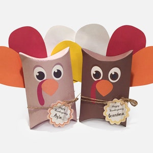 Puede incluir: Dos cajas de almohada marrones y grises decoradas como pavos con plumas de papel naranja, amarillo y blanco. Cada caja tiene una etiqueta con el texto "Feliz Día de Acción de Gracias" y un nombre, "Ayla" y "Abuela".
