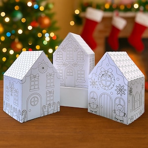May include: Three white paper gingerbread houses with black outlines, ready to be colored. Each house features different Christmas-themed designs, including candy canes, gingerbread people, and wreaths. The houses are set on a wooden surface with a blurred Christmas tree and stockings in the background.