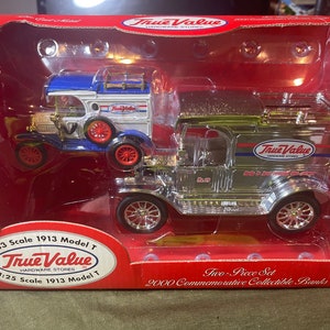 May include: Two vintage-style metal toy banks shaped like 1913 Model T Ford trucks. One bank is silver with a red and blue True Value Hardware Stores logo. The other bank is blue, white, and red with a True Value Hardware Stores logo. The banks are displayed in a red box with the text "Two-Piece Set 2000 Commemorative Collectible Banks".