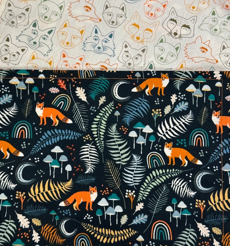 May include: Two fabric patterns are shown. The top fabric has a repeating pattern of fox faces in orange, blue, and brown on a cream background. The bottom fabric has a dark blue background with foxes, mushrooms, rainbows, ferns, and moons.