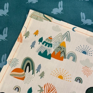 May include: A white fabric with a colorful mountain, rainbow, and tree pattern. The fabric is bordered by a teal fabric with a repeating rabbit silhouette design. The mountain design includes teal, orange, and gray colors.