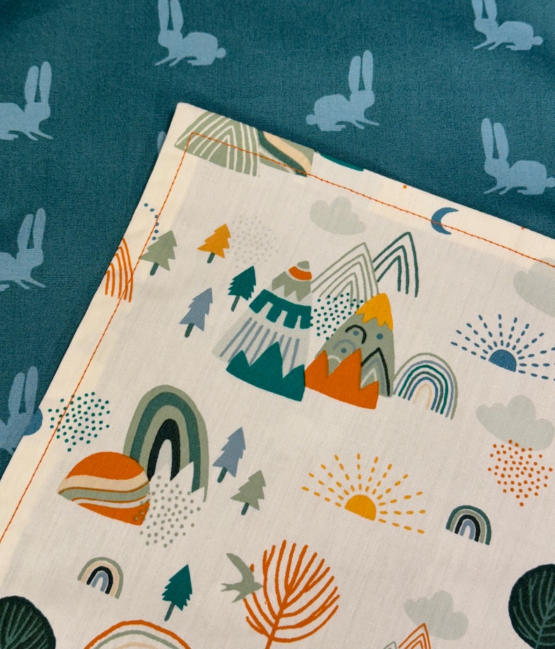 May include: A white fabric with a colorful mountain, rainbow, and tree pattern. The fabric is bordered by a teal fabric with a repeating rabbit silhouette design. The mountain design includes teal, orange, and gray colors.