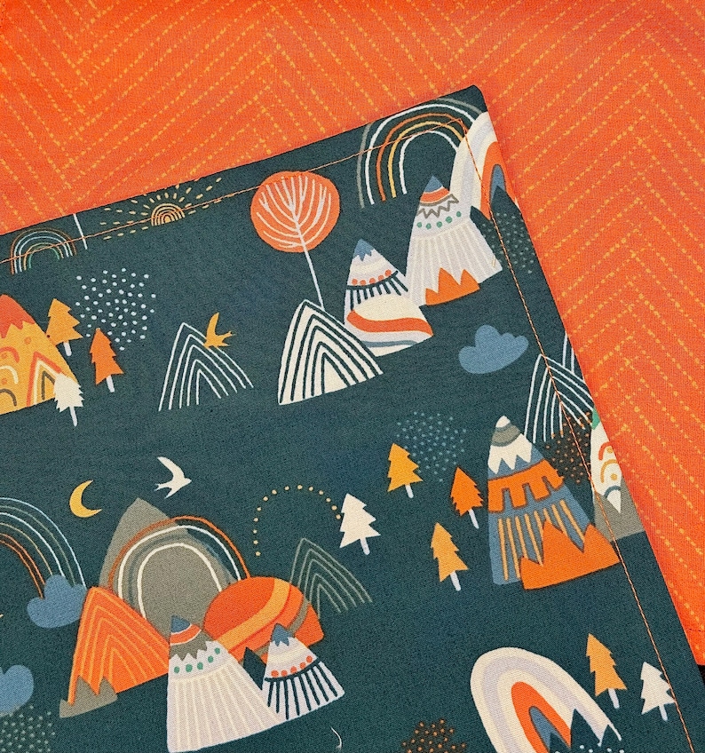 May include: A dark teal fabric with a mountain and rainbow pattern. The design includes orange, white, and gray mountains, rainbows, trees, and a crescent moon. The background is a solid orange fabric with a subtle chevron pattern.