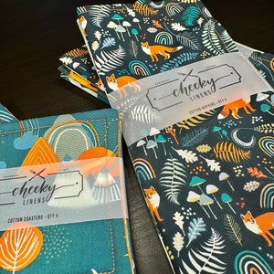May include: A set of four cotton napkins and a set of four cotton coasters. The napkins and coasters feature a dark teal background with a repeating pattern of orange foxes, mushrooms, ferns, and rainbows. The text "cheeky linens" is visible.
