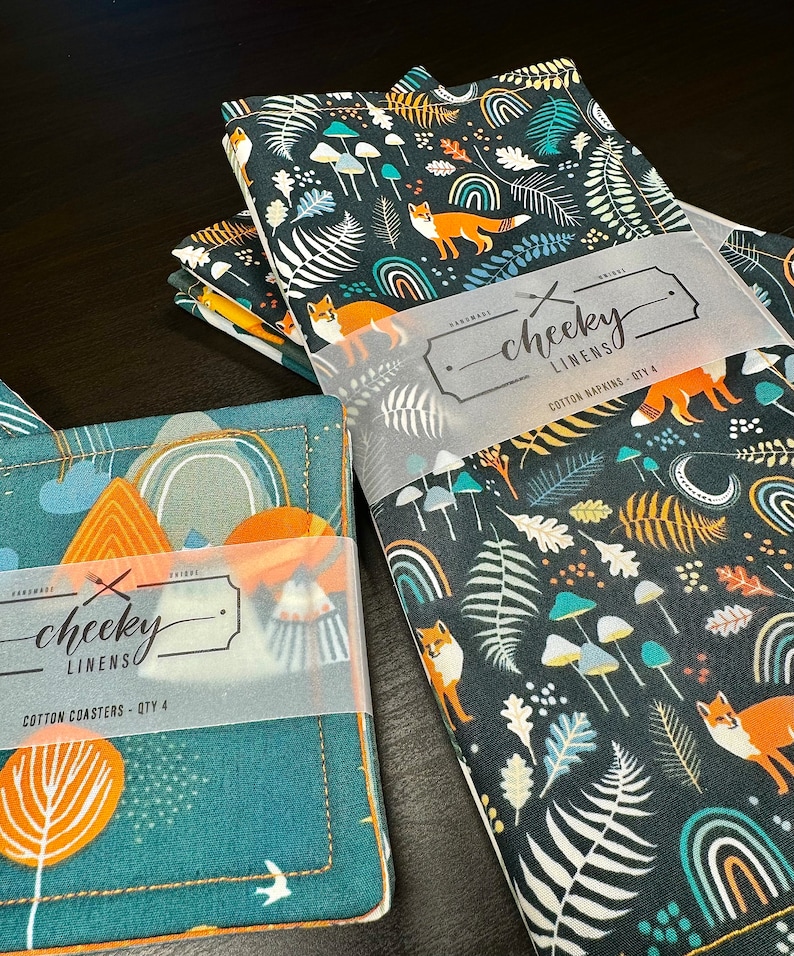 May include: A set of four cotton napkins and a set of four cotton coasters. The napkins and coasters feature a dark teal background with a repeating pattern of orange foxes, mushrooms, ferns, and rainbows. The text "cheeky linens" is visible.