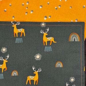 May include: Two fabric patterns are shown. The top fabric is mustard yellow with a pattern of white acorns. The bottom fabric is dark gray with a pattern of yellow deer, rainbows, and suns. The deer have large antlers.