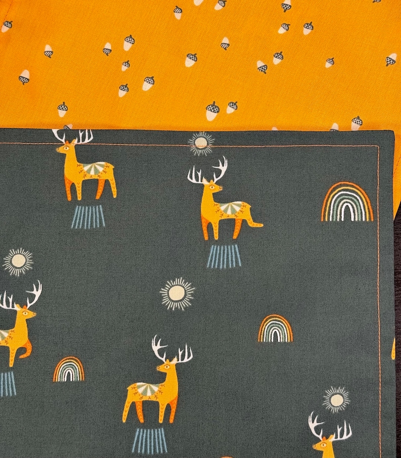 May include: Two fabric patterns are shown. The top fabric is mustard yellow with a pattern of white acorns. The bottom fabric is dark gray with a pattern of yellow deer, rainbows, and suns. The deer have large antlers.