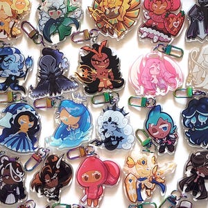 May include: A collection of twenty-four acrylic keychains featuring various cartoon character illustrations with diverse color palettes including shades of blue, red, gold, black, and pink. Each keychain is attached to a small metal lobster clasp.
